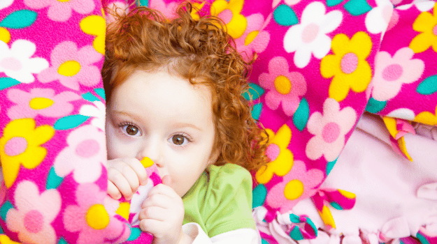 Red headed child with curly hair hiding behind a pink floral tied blanket